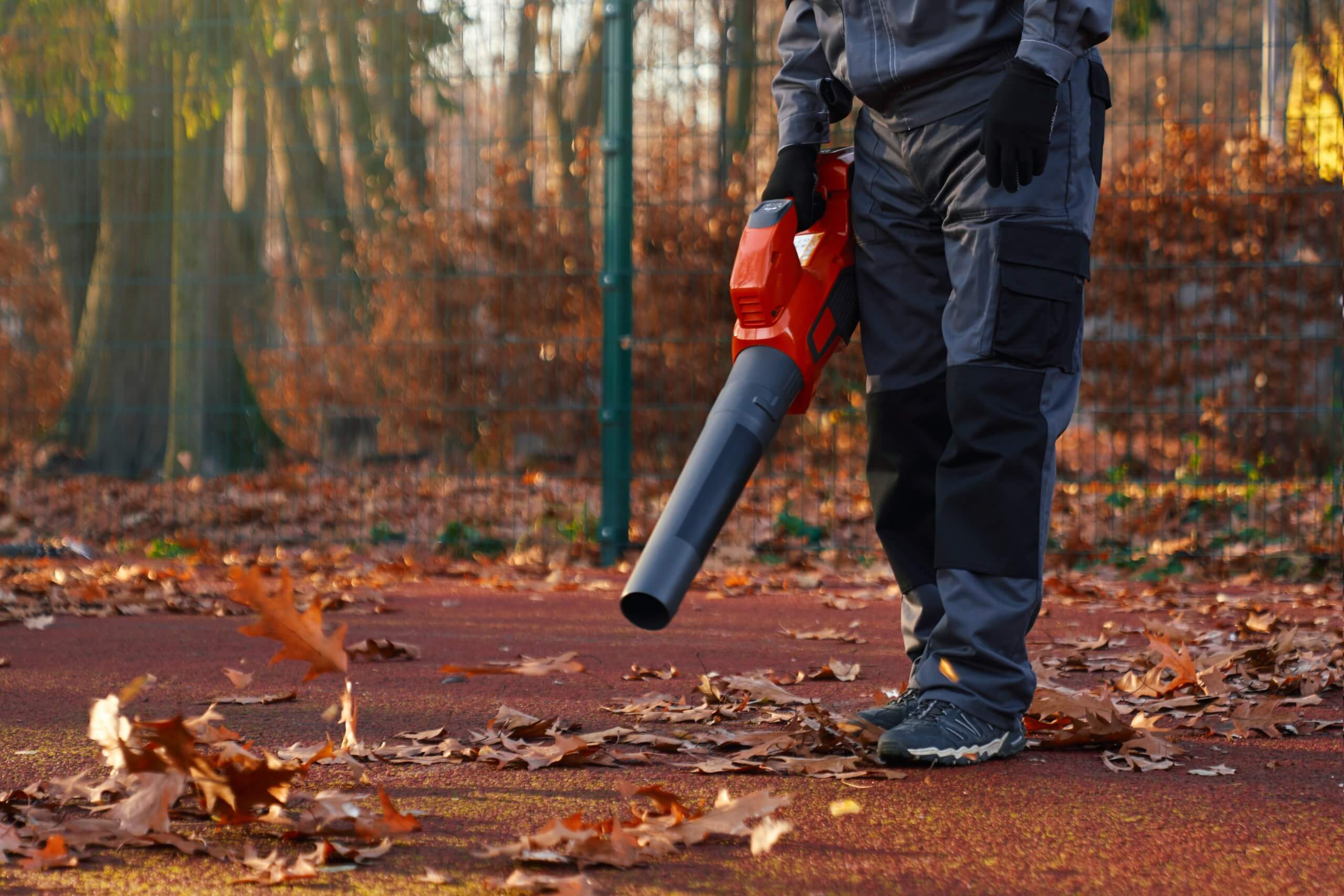 Unrecognizable maintenance man wearing blue uniform and gloves, working with portable leaf blower in morning. Cropped view of male worker holding, using leaf blower at park area. Concept of work.